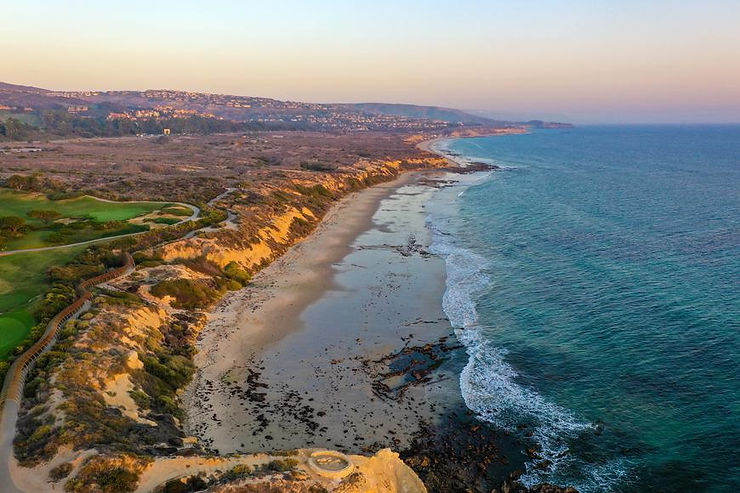 Aerial view of a scenic coastline with cliffs, sandy beach, and blue ocean waves at sunset.