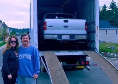 Couple standing in front of a moving truck as their Ford pickup is loaded via ramp.