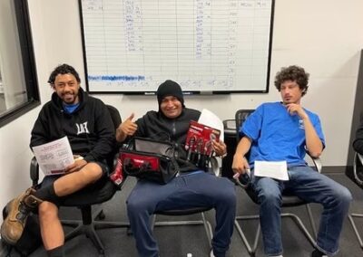 Three employees seated in office chairs, smiling and holding tools in front of a performance tracking whiteboard.